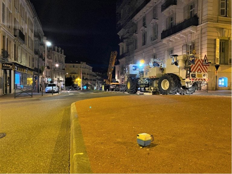 A SmartSolo node seismometer sits on a street at night during a seismic survey with a large vibrator truck in the background.