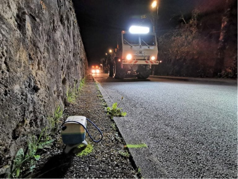 A SmartSolo node seismometer is placed on the ground next to a road at night during a seismic survey with a vibrator truck in the background.