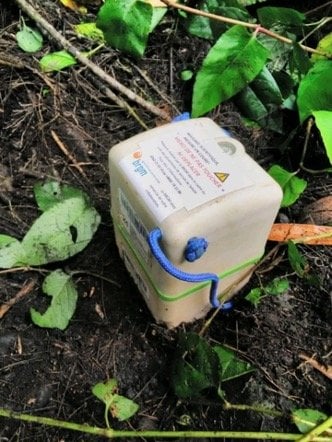 A beige SmartSolo node with blue handles sits on the ground surrounded by green leaves and brown dirt, used for seismic activity monitoring.