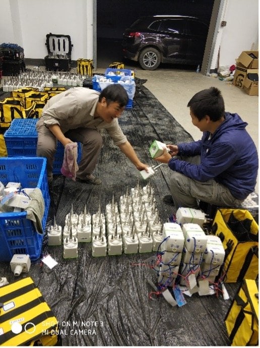 Two men prepare SmartSolo nodes and seismometers for geophysical exploration, surrounded by equipment bags and crates.