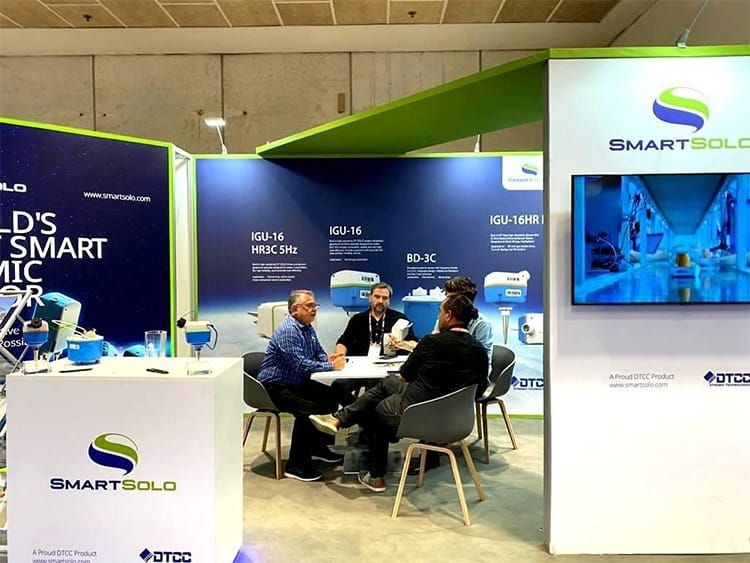 Four people discussing SmartSolo seismic nodes and seismometers at a trade show booth for geophysical exploration.