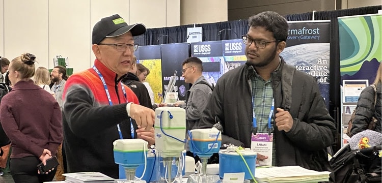 Two men discuss SmartSolo seismometers and nodes for seismic surveys and earthquake monitoring at a trade show booth.