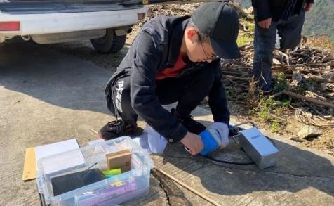 A person in a black jacket and cap is crouching, connecting a blue cable to a SmartSolo seismometer node on the ground. A white vehicle is parked behind them. This is part of a seismic survey for geophysical exploration.