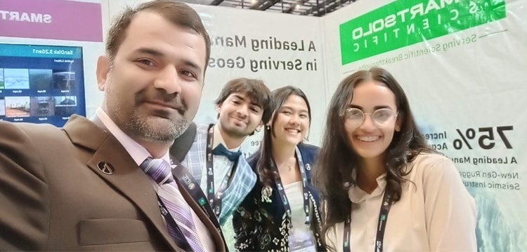 Four people smiling at the SmartSolo Scientific booth. Text on banner: "SMARTSOLO SCIENTIFIC Serving Scientific Breakthroughs A Leading Manufacturer in Serving Geophysical Exploration".