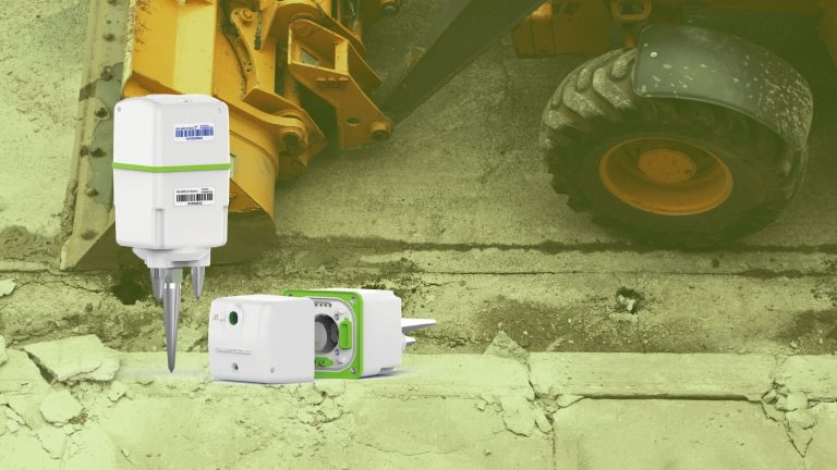 Two SmartSolo nodes, a type of seismometer, are shown in the foreground with a large yellow excavator in the background. These nodes are used for seismic survey and earthquake monitoring.