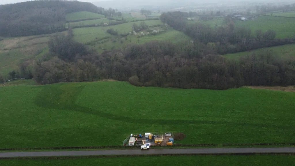 Aerial view of a green field with a cluster of geophysical exploration equipment and vehicles near a road, with hills and trees in the background.