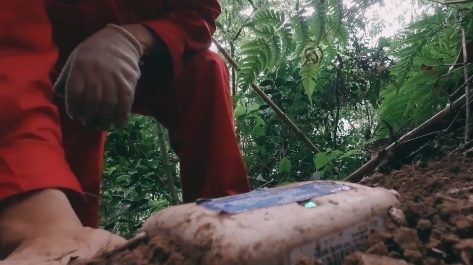 Person in red suit and gloves placing a SmartSolo node in the ground for seismic activity monitoring.