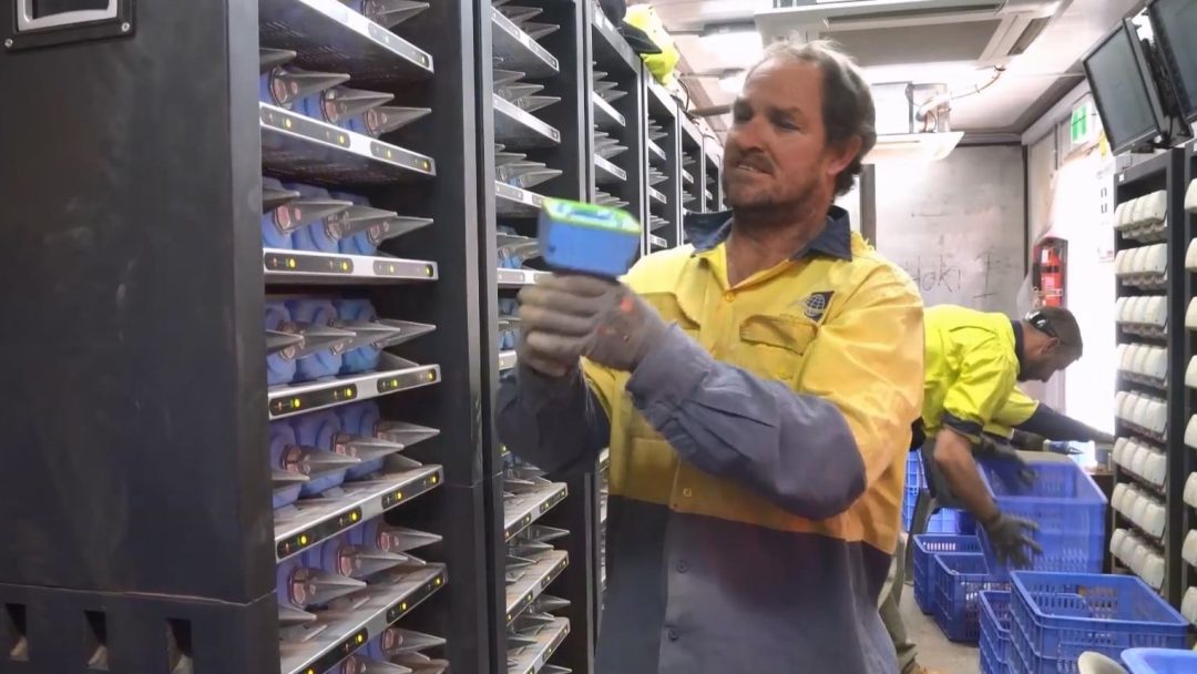 A man in a yellow and blue work shirt holds a battery part of seismic node before a charging rack. Another man works in the background.