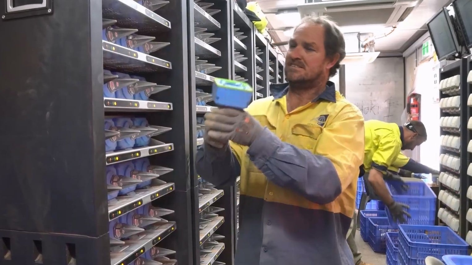 Workers in a room with racks of SmartSolo nodes, one scanning a node and another placing nodes battery into crates for charging.