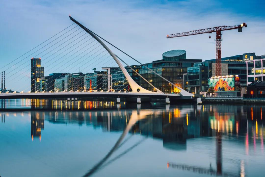 Modern bridge and buildings reflected in the water at dusk, with a large crane in the background.