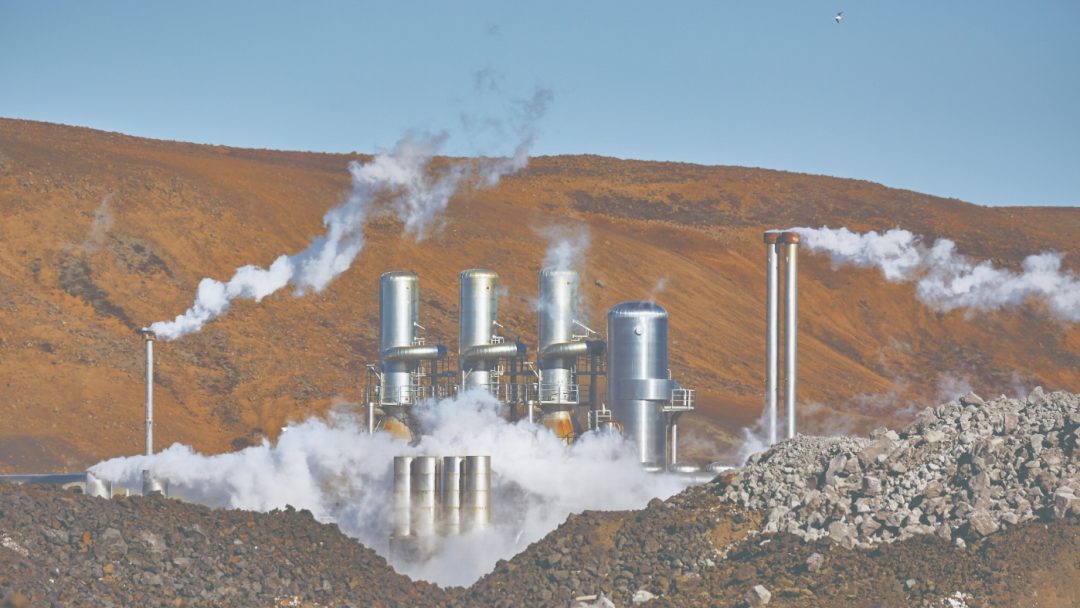 Geothermal power plant with steam vents and industrial equipment set against a barren, rocky landscape under a clear blue sky