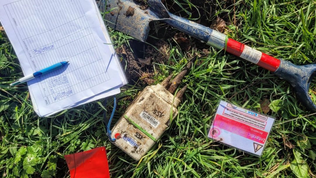 A seismometer IGU-16HR 3C on grass with a shovel, clipboard, pen, and a warning card, used for earthquake monitoring and geophysical exploration.