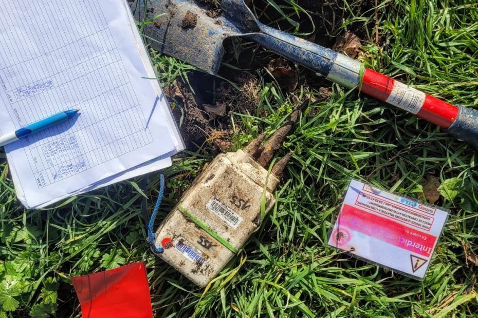 A seismometer IGU-16HR 3C on grass with a shovel, clipboard, pen, and a warning card, used for earthquake monitoring and geophysical exploration.