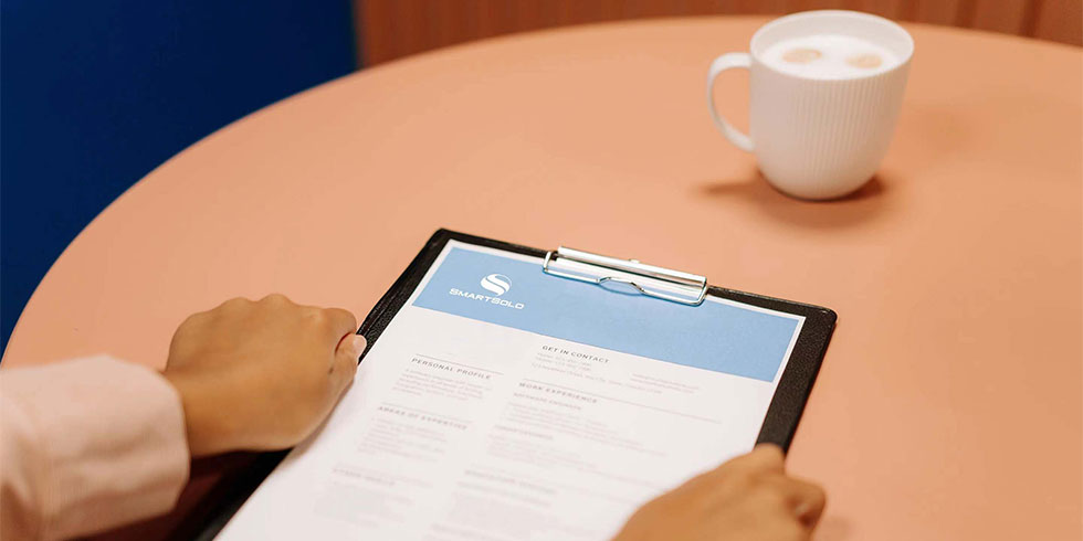 Hands holding a SmartSolo document on a clipboard. Coffee cup on the table. Document includes personal profile and work experience.